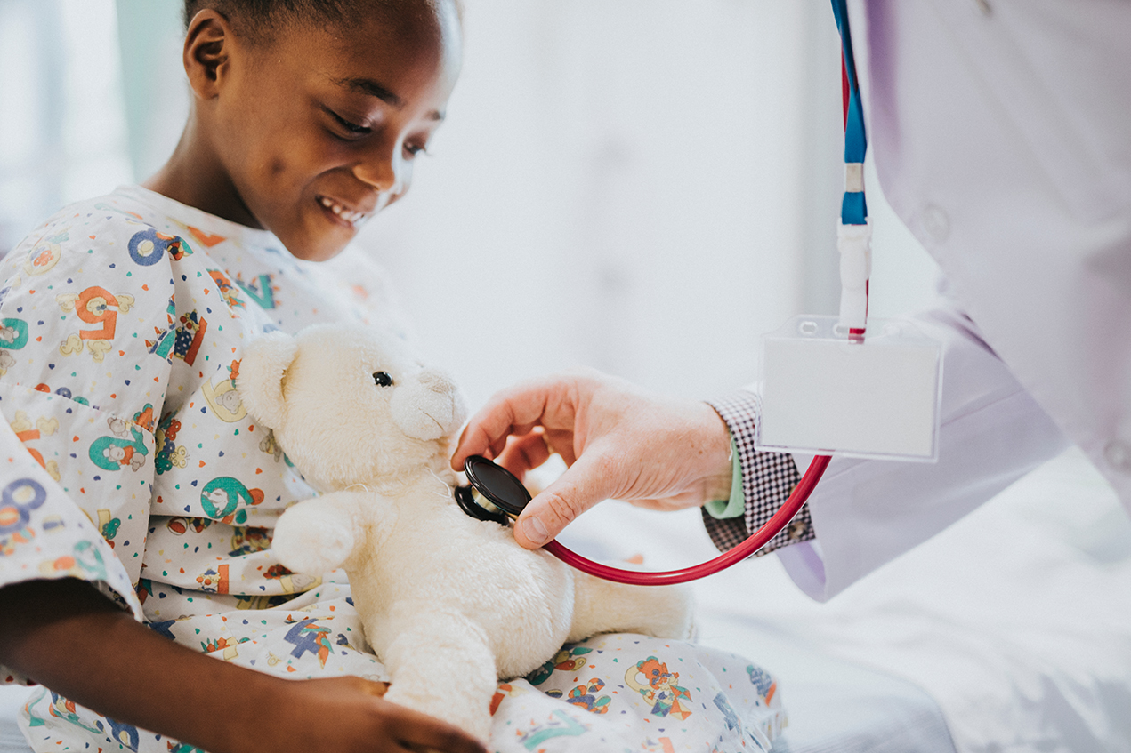 A child in a hospital gown smiles as a doctor holds a stethoscope to her teddy bear's chest